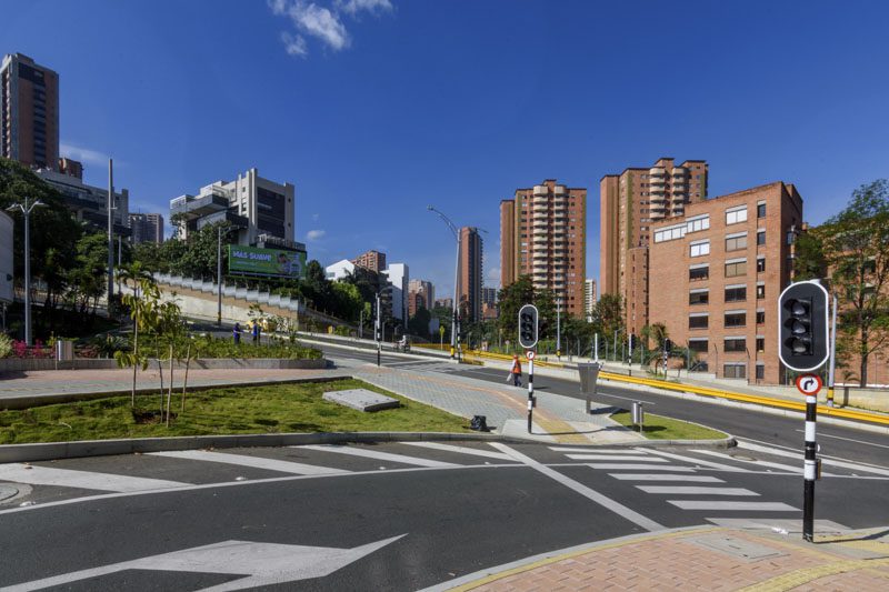 Intersección con semáforos, áreas verdes y edificios modernos bajo cielo azul despejado.